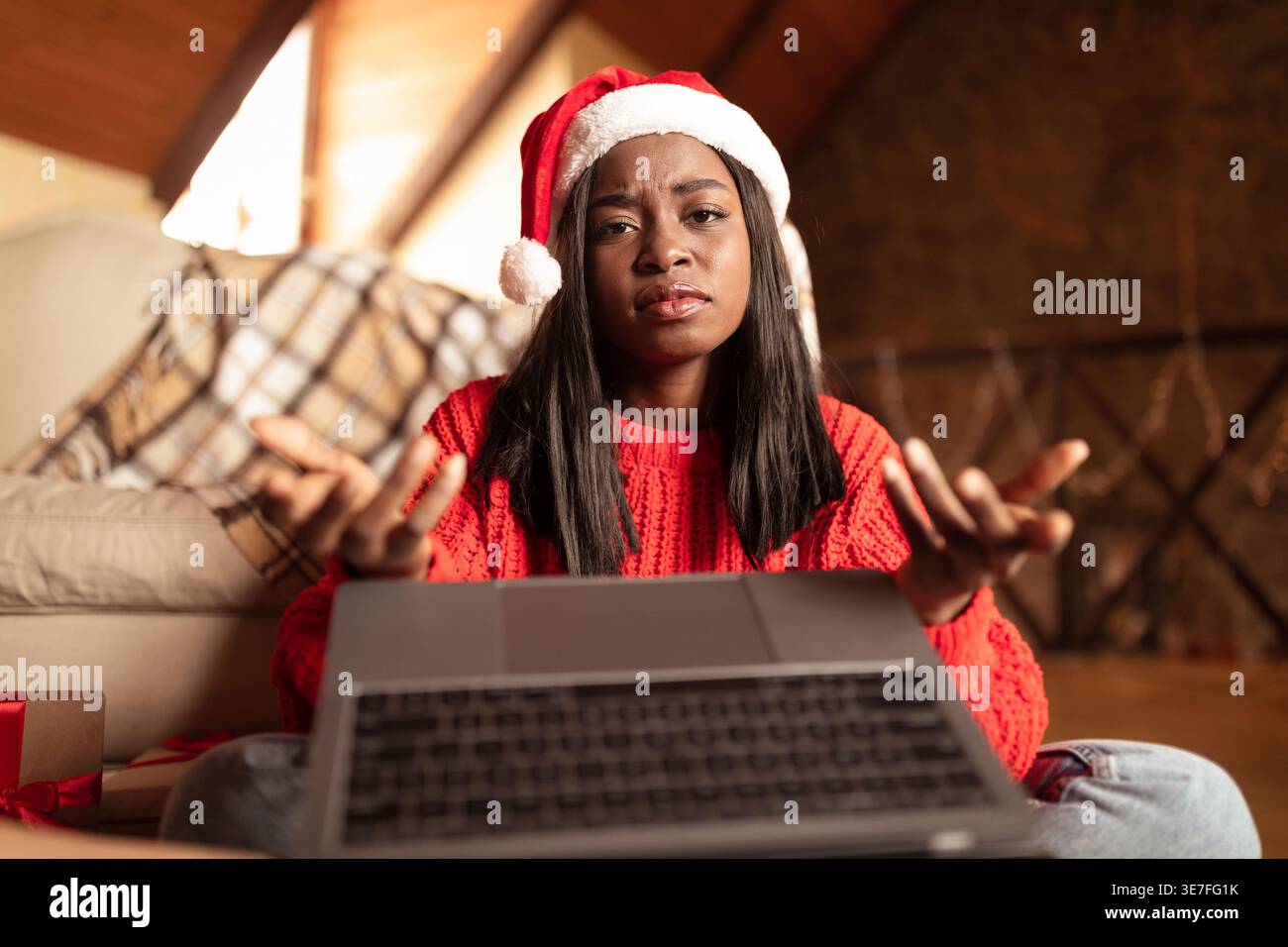 Bouleversée adolescente noire portant un chapeau de Père Noël faisant des gestes des mains tout en ayant un appel vidéo à l'aide d'un ordinateur portable assis sur le sol dans le salon à la maison Banque D'Images