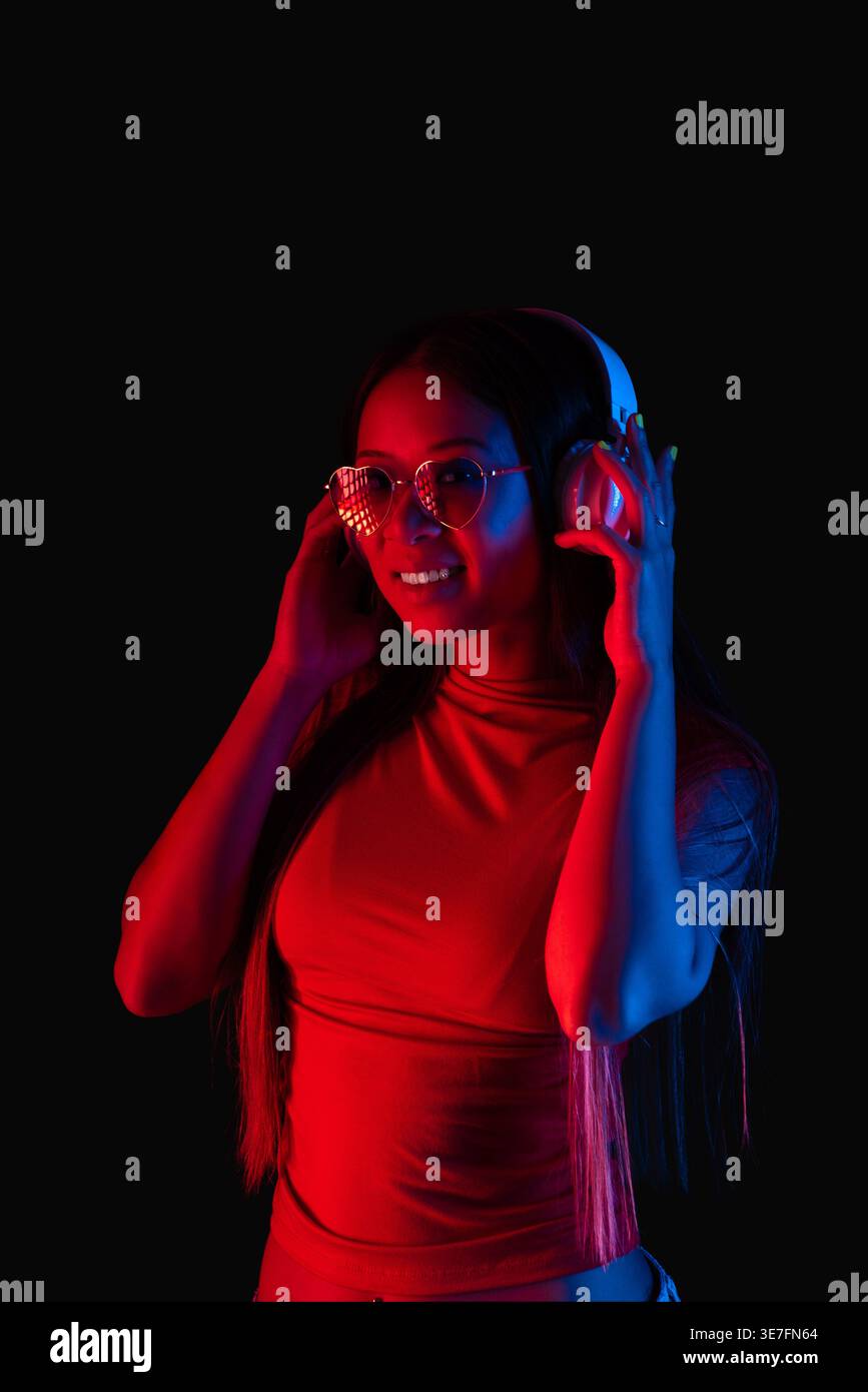 Femme asiatique avec des lunettes de soleil en forme de coeur écoutant de la musique sur des écouteurs debout sur fond noir avec lumière rouge en studio Banque D'Images
