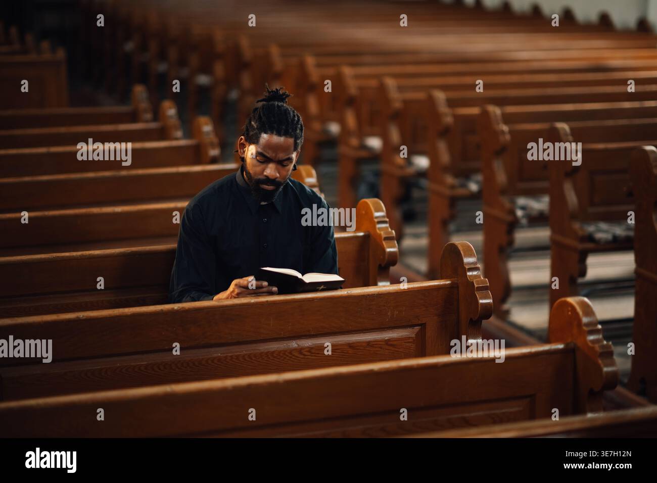 Prêtre noir avec moustache et barbe lisant la Bible tout en étant assis sur un banc en bois dans l'église Banque D'Images