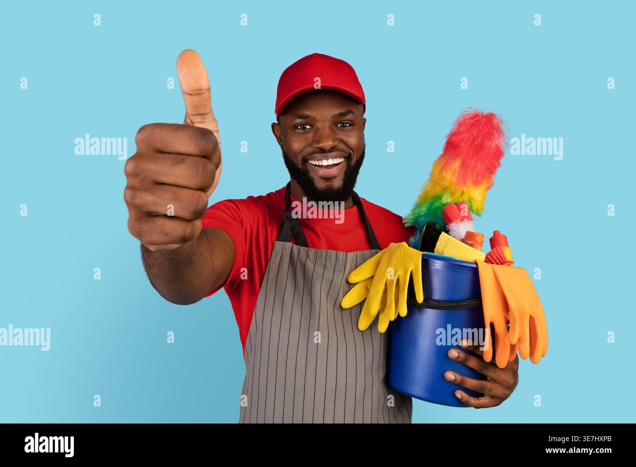 Chapeau de port et tablier noir nettoyant masculin montrant les pouces vers le haut et tenant le panier d'outils de nettoyage debout sur fond bleu dans le studio Banque D'Images