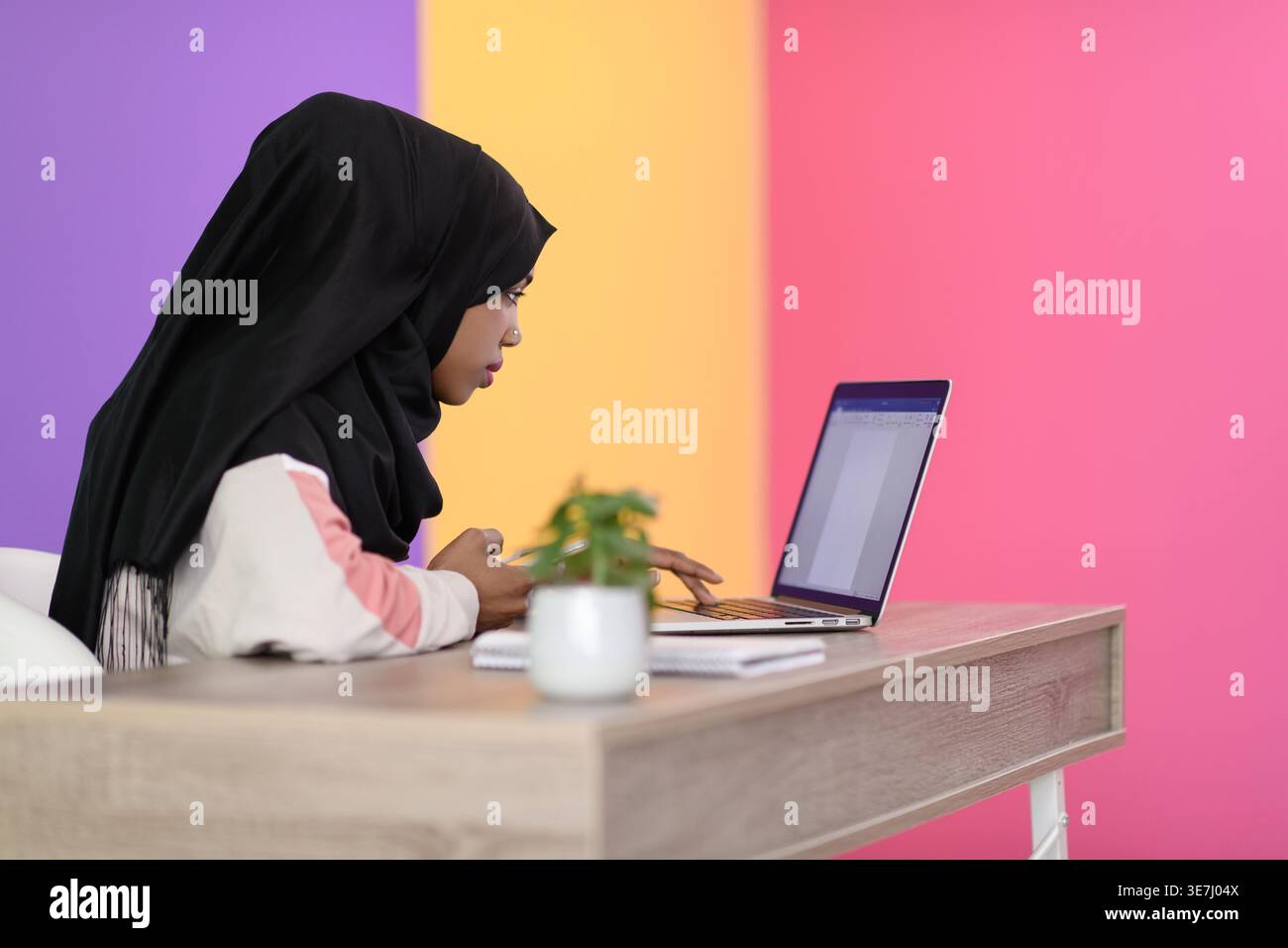 Femme d'affaires musulmane noire sérieuse avec perçage du nez portant le hijab travaillant sur ordinateur portable tout en étant assise à table dans le bureau Banque D'Images