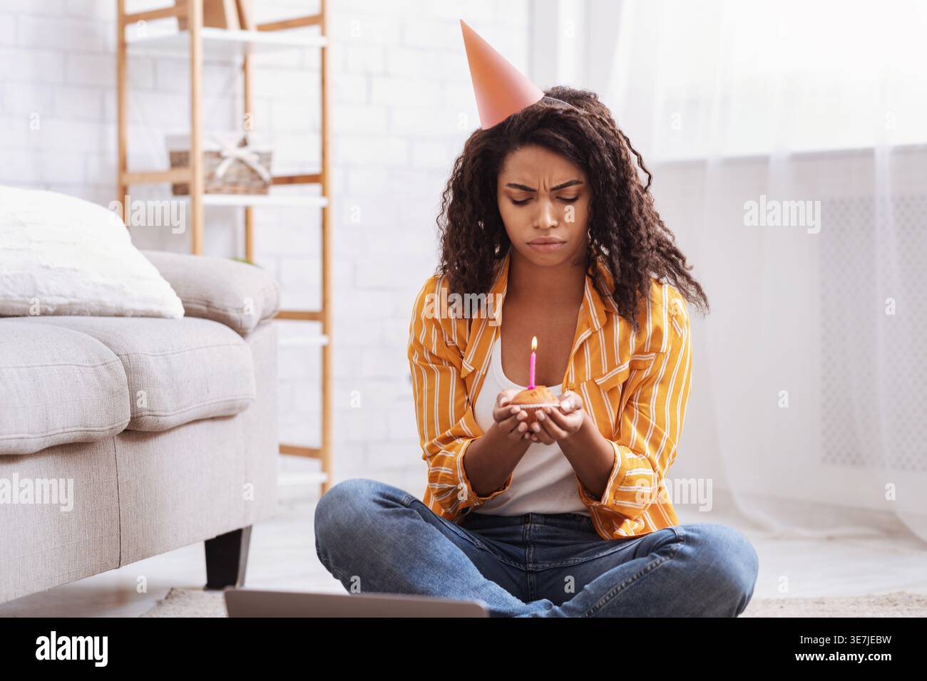 Contrarié femme noire portant chapeau de fête tenant cupcake avec bougie tout en étant assis sur le tapis de sol avec ordinateur portable dans le salon à la maison Banque D'Images