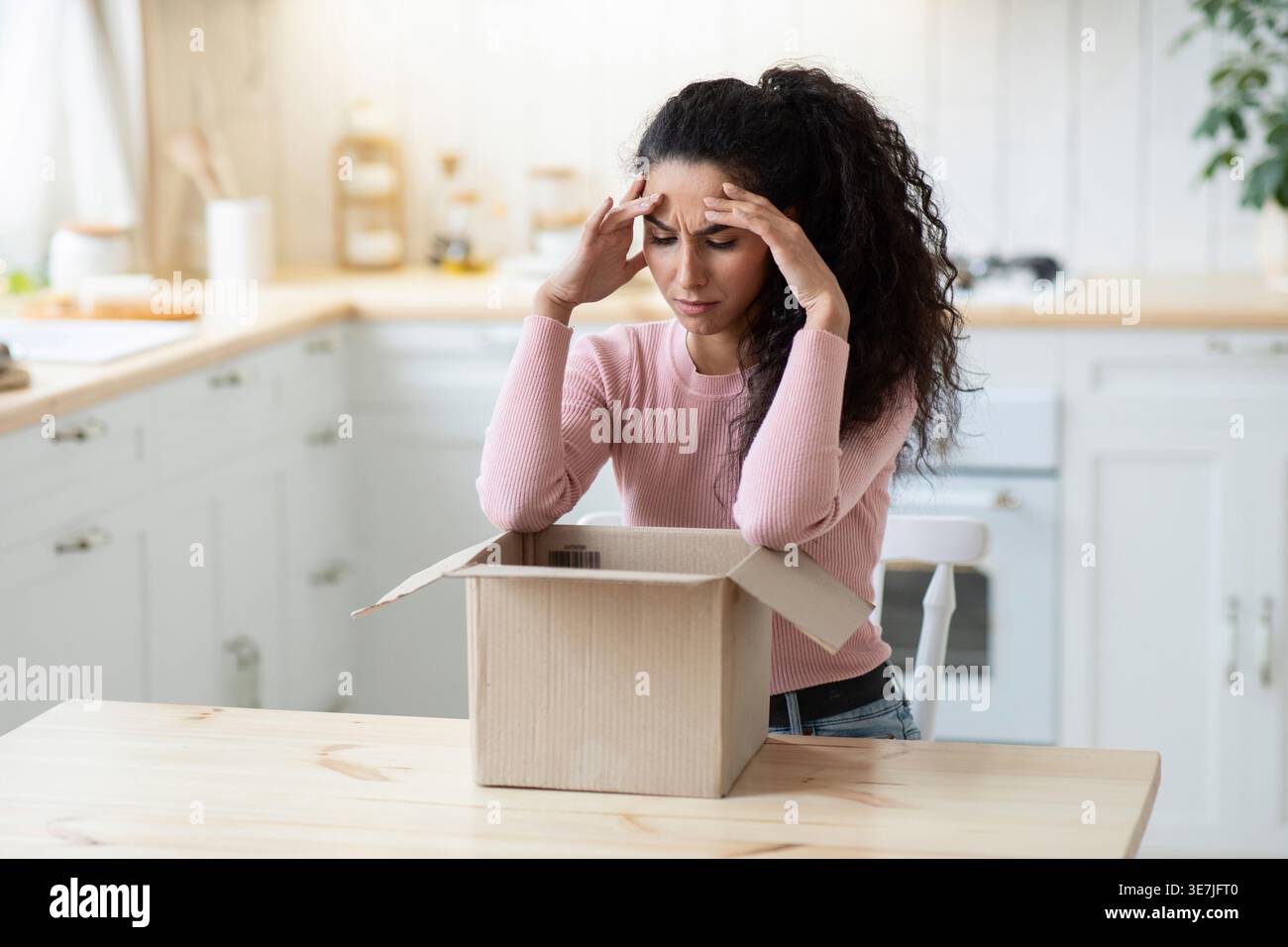 Femme frustrée du moyen-Orient touchant la tête regardant la boîte en carton ayant un problème avec son paquet tout en étant assise à table dans la cuisine à la maison Banque D'Images