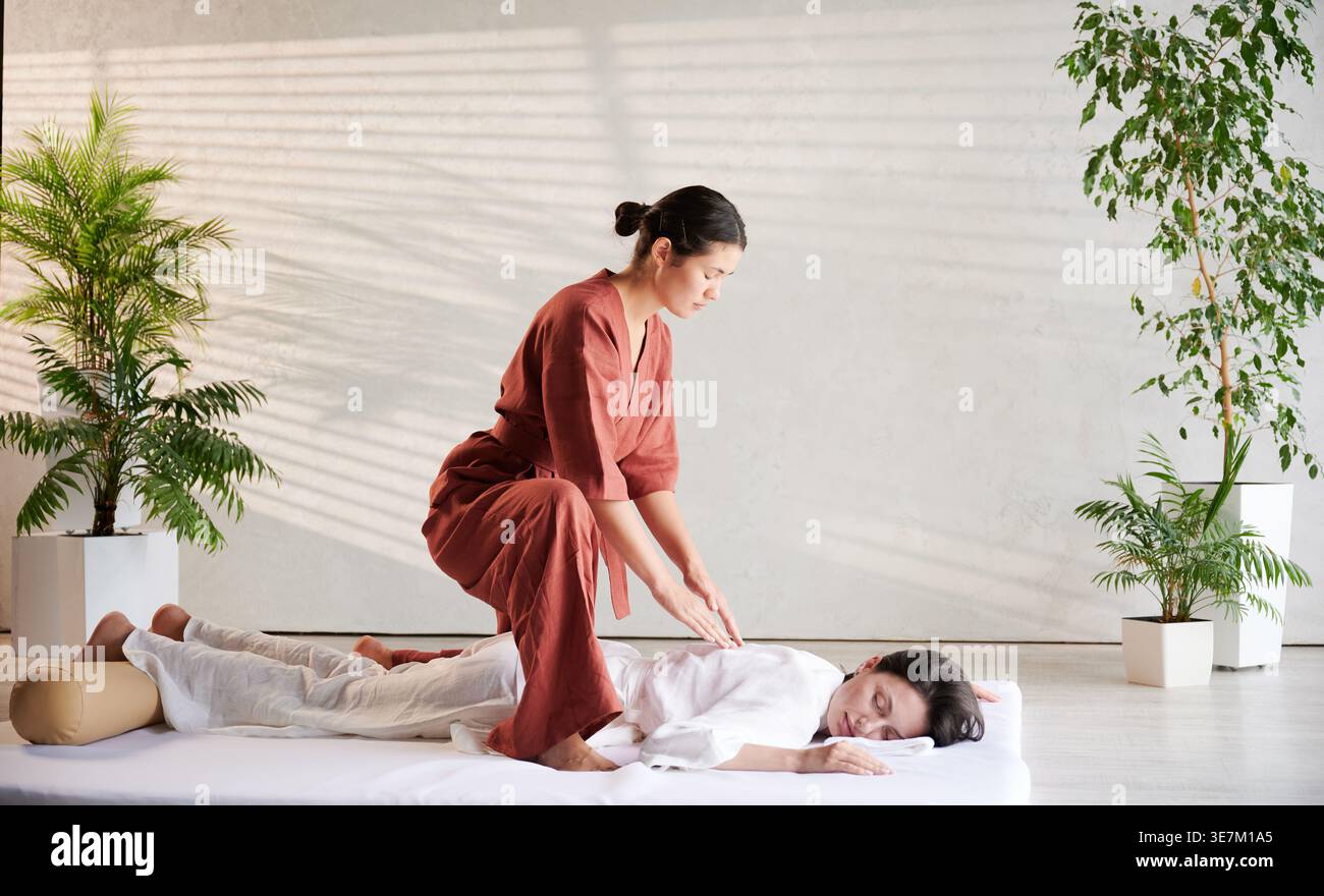 Masseuse faisant massage yoga thaïlandais. Thérapeute de femme faisant le traitement de massage traditionnel pour la patiente féminine à l'intérieur. Client allongé sur tapis, méditant, profitant d'un massage des mains dans une atmosphère calme. Banque D'Images