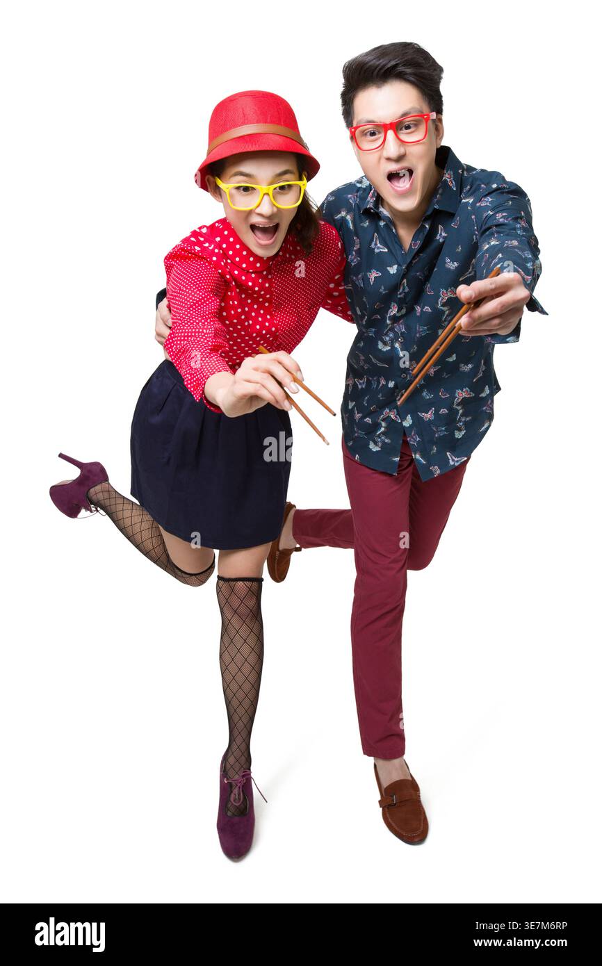 Joyeux couple chinois obtenant quelque chose en utilisant des baguettes tout en se tenant debout ensemble sur un pied sur fond blanc en studio Banque D'Images