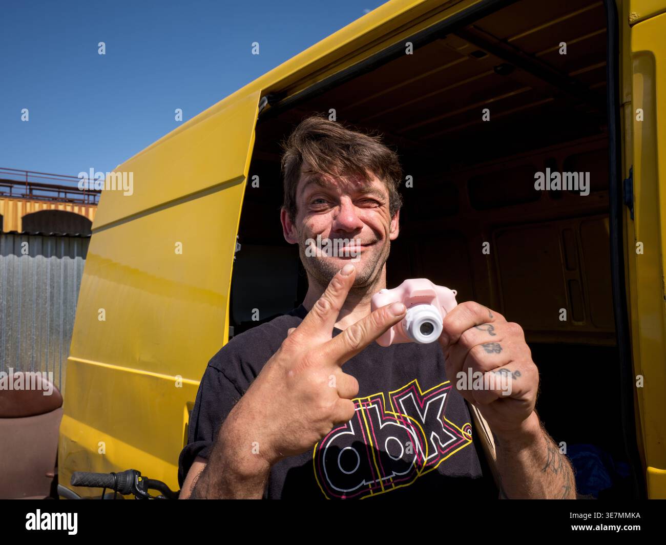 Homme souriant montrant un signe de victoire et tenant une petite caméra devant un van jaune à un marché aux puces de Prague Banque D'Images