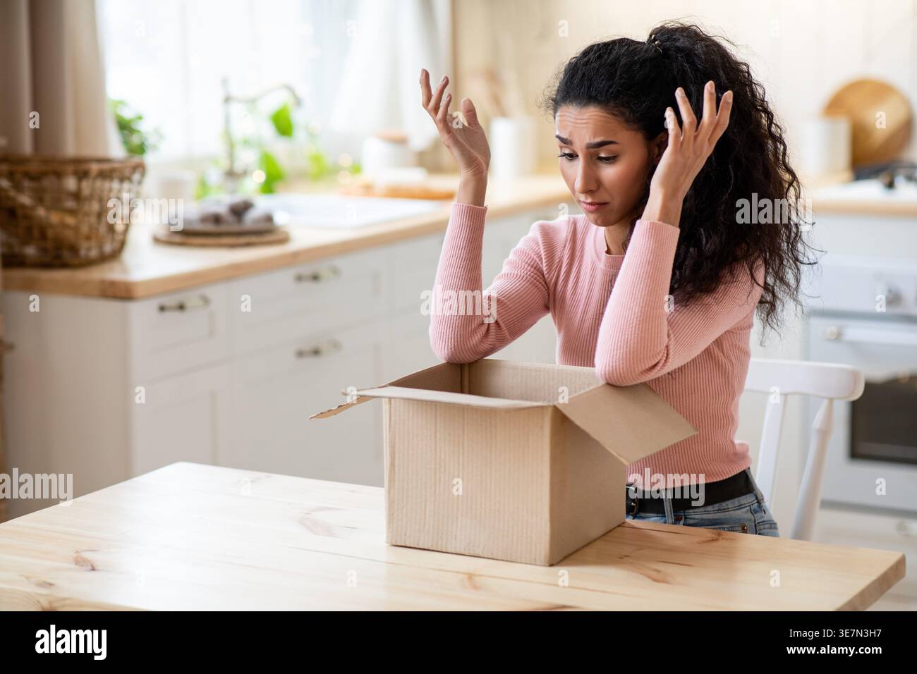 Bouleversée femme du moyen-Orient faisant des gestes des mains regardant la boîte en carton ayant un problème avec son paquet tout en étant assis à table dans la cuisine à la maison Banque D'Images