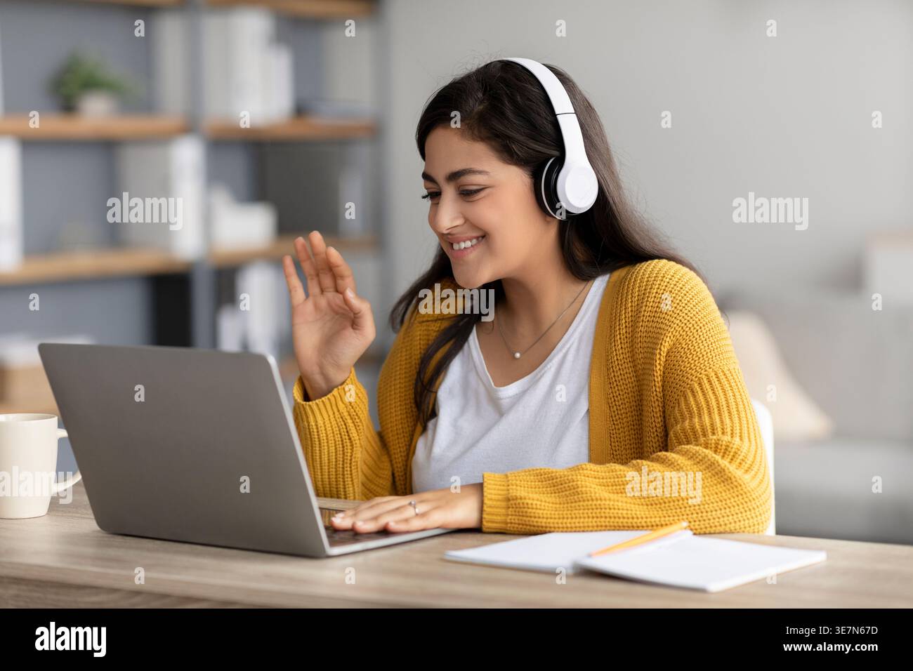 Femme du moyen-Orient souriante portant des écouteurs agitant la main et ayant un appel vidéo sur ordinateur portable tout en étant assis au bureau avec ordinateur portable et tasse de café je... Banque D'Images