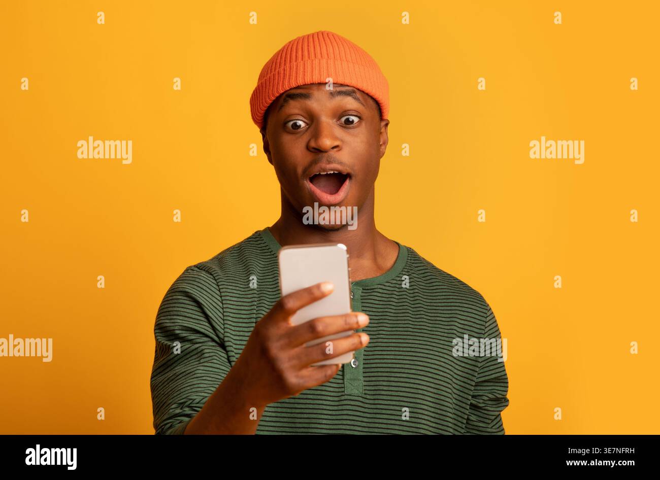 Homme noir portant un bonnet avec une expression choquée à l'aide d'un smartphone tout en se tenant sur fond jaune dans le studio Banque D'Images