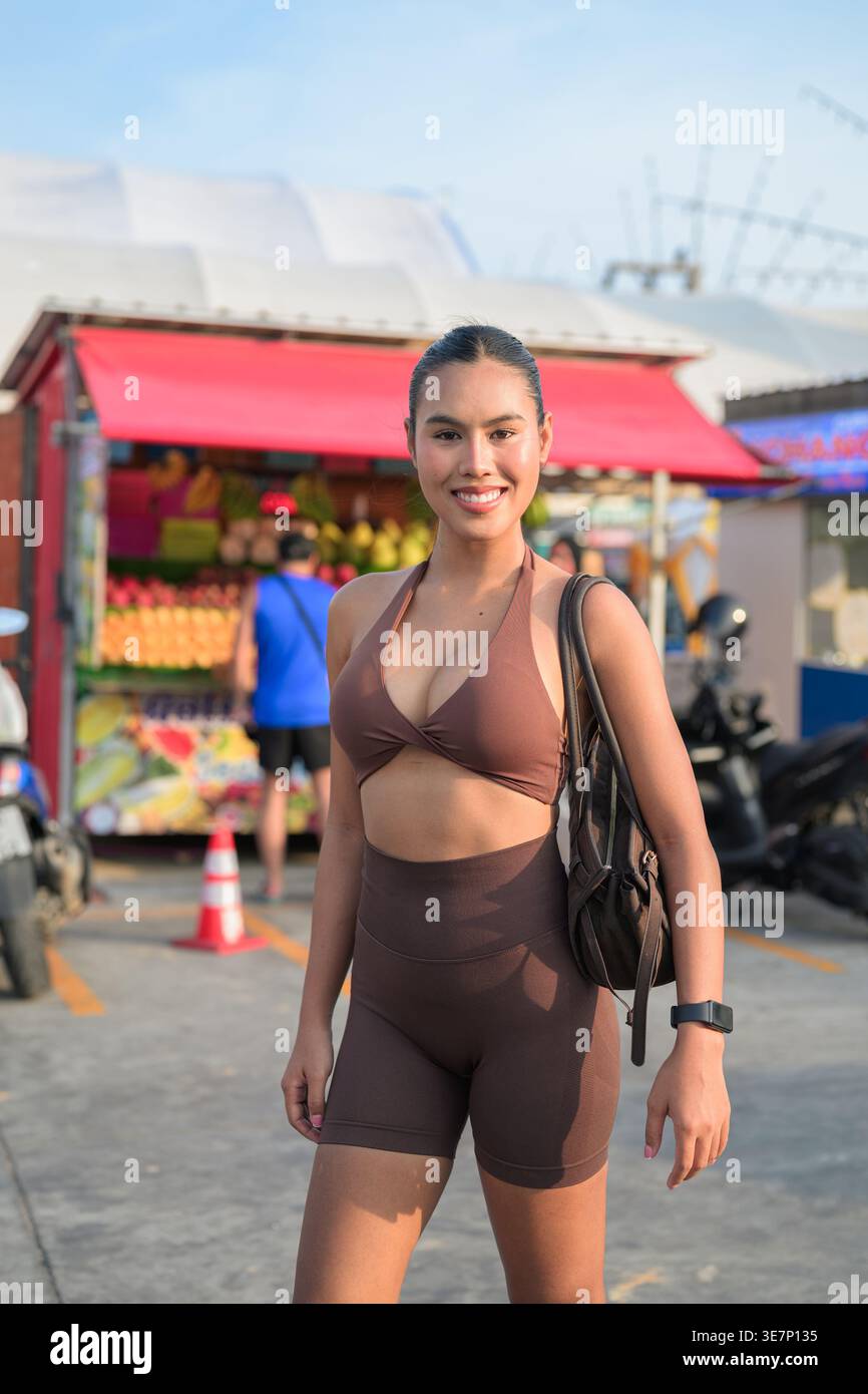 Une jeune femme confiante en vêtements de sport côtelés bruns sourit tout en se tenant devant un étal coloré de fruits tropicaux à un marché de rue en plein air. Banque D'Images