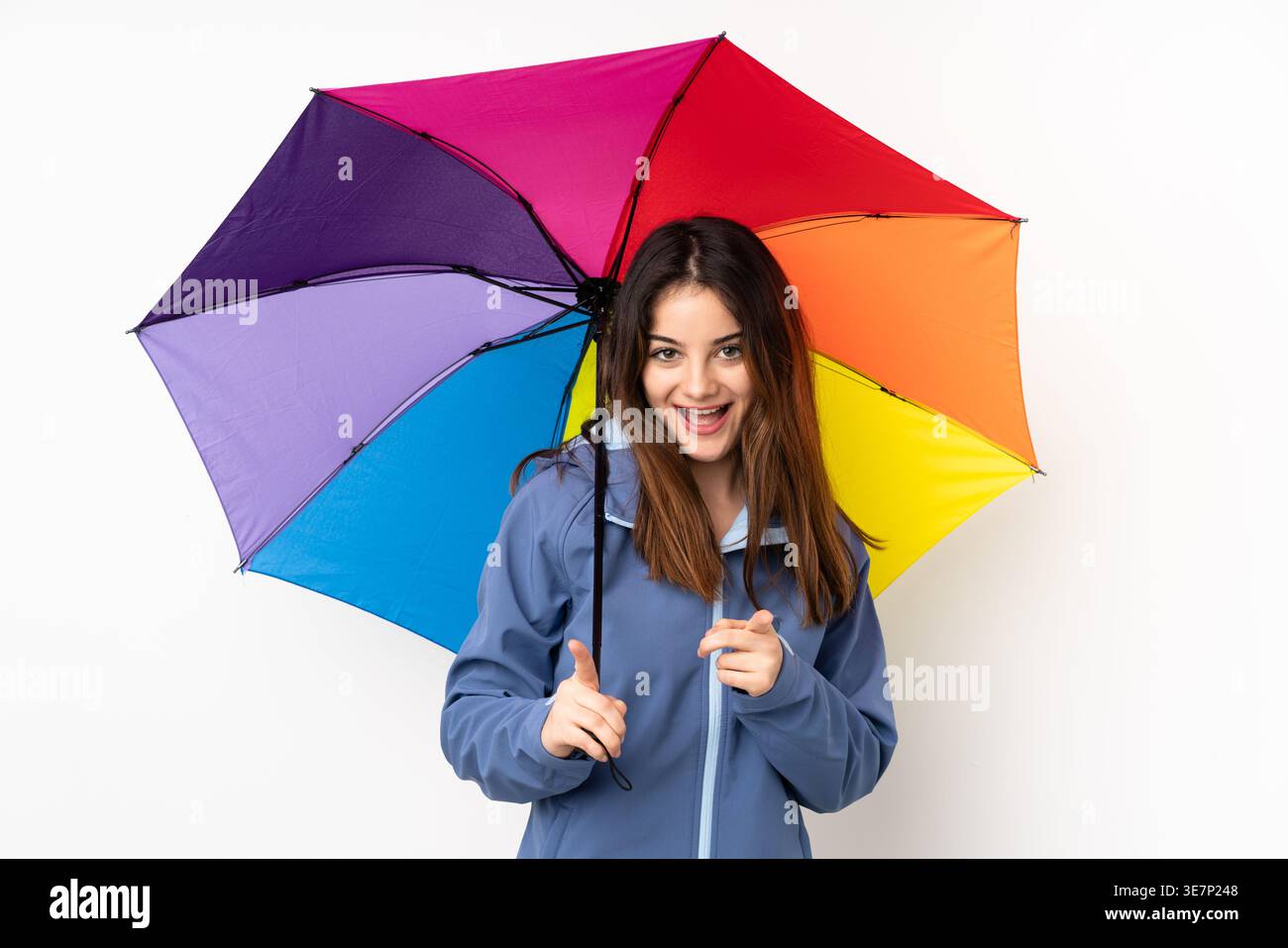 Femme hispanique portant une veste tenant un parapluie arc-en-ciel et pointant le doigt vers l'avant tout en se tenant debout sur fond blanc dans le studio Banque D'Images
