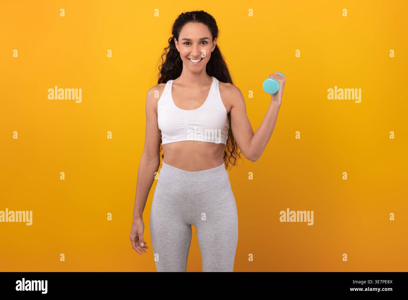 Femme du moyen-Orient souriante avec des cheveux bouclés portant des vêtements de sport tenant un haltère debout sur fond jaune dans le studio Banque D'Images