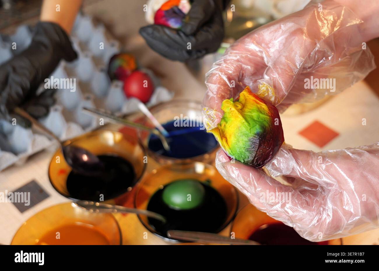 Les personnes portant des gants teignent les œufs de Pâques à l'aide de liquides colorés, avec les mains tenant des œufs multicolores et des bols de colorants bleus et foncés sur une table de fête Banque D'Images