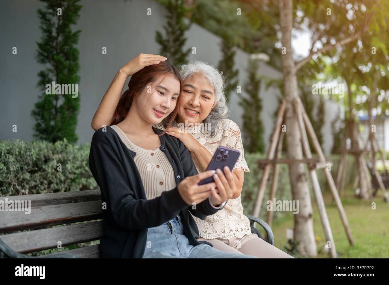 Femme thaïlandaise ayant du plaisir à partager smartphone avec sa grand-mère tout en étant assis sur un banc en bois dans le parc pendant la journée Banque D'Images