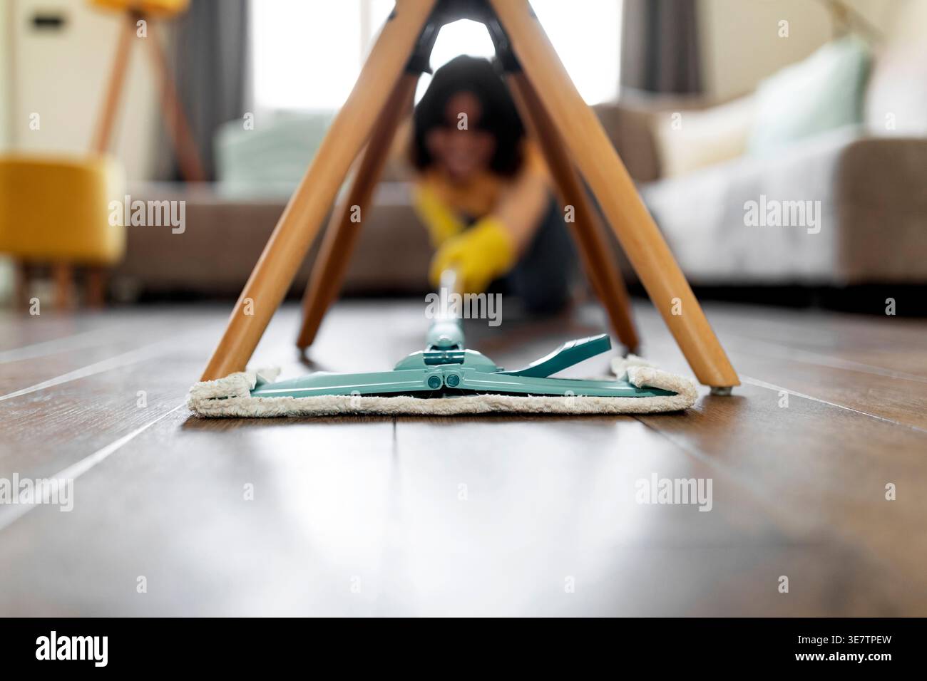 Adolescente hispanique portant des gants en caoutchouc à l'aide d'une vadrouille dans le nettoyage du plancher en bois sous la chaise dans le salon à la maison Banque D'Images