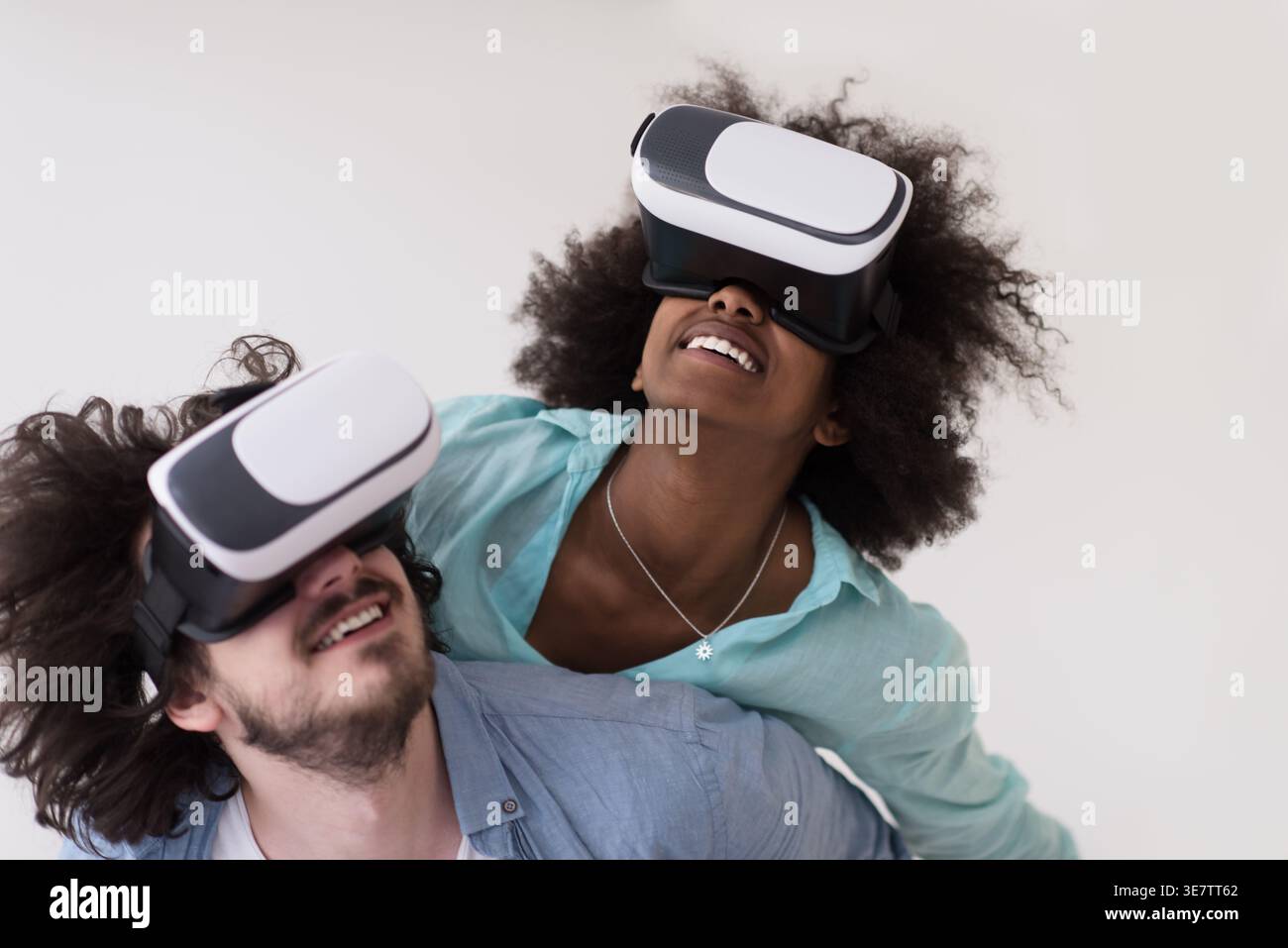 Couple noir et caucasien portant des casques VR ayant un plaisir piggyback équitation et expérimentant la réalité virtuelle sur fond blanc en studio Banque D'Images
