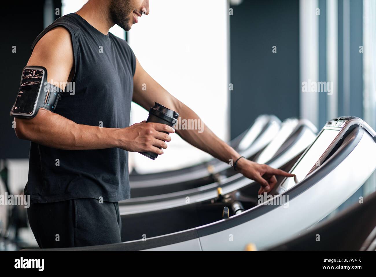 Image recadrée d'un homme noir avec une barbe portant un brassard tenant une bouteille shaker et sélectionnant le mode sur le tapis roulant avant de faire du jogging dans la salle de gym Banque D'Images