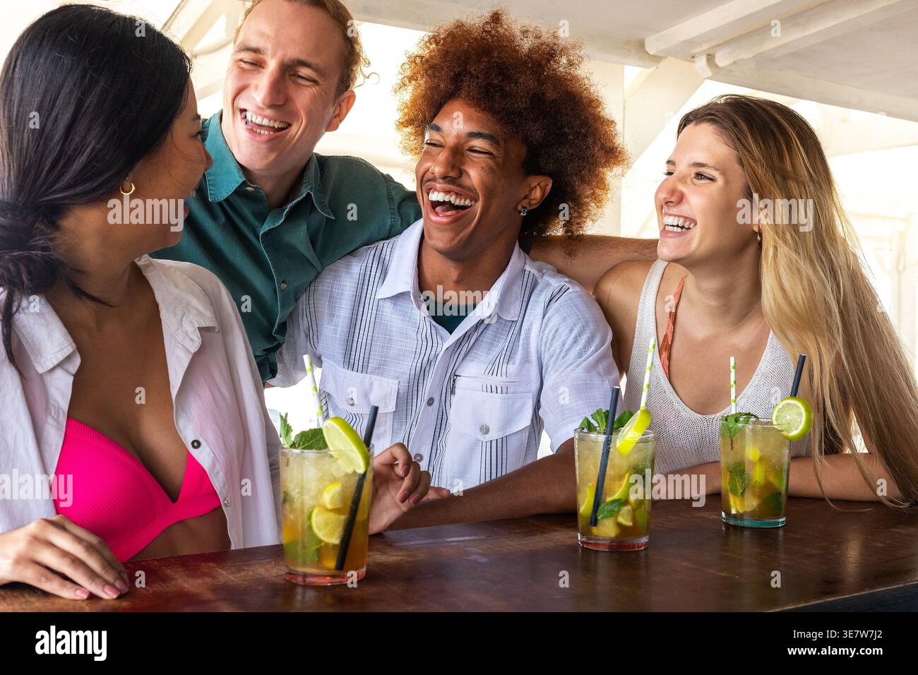 Homme noir joyeux avec rire afro tout en se tenant debout au comptoir avec divers amis prenant des cocktails mojito au bar de la plage pendant les vacances d'été Banque D'Images