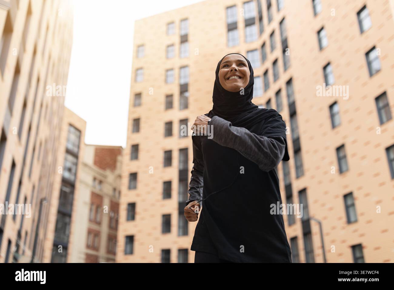 Femme musulmane du moyen-Orient souriante avec tracker de fitness portant hijab et vêtements de sport courant dans la rue pendant la journée Banque D'Images
