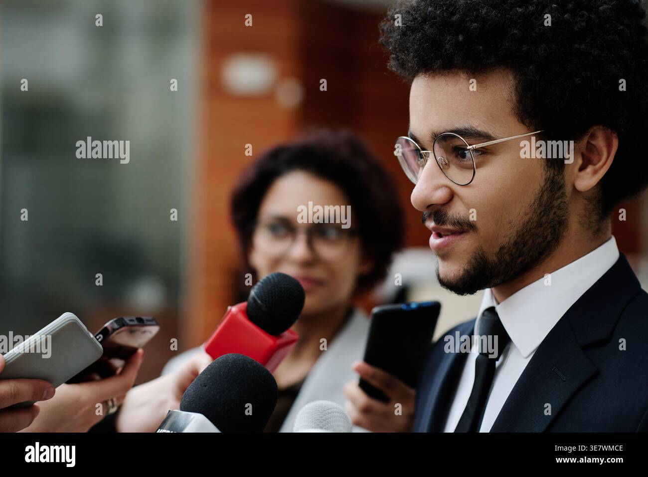 Homme d'affaires de race mixte en costume et lunettes parlant aux journalistes donnant une interview à la conférence d'affaires tout en se tenant debout à l'extérieur du bureau bui... Banque D'Images
