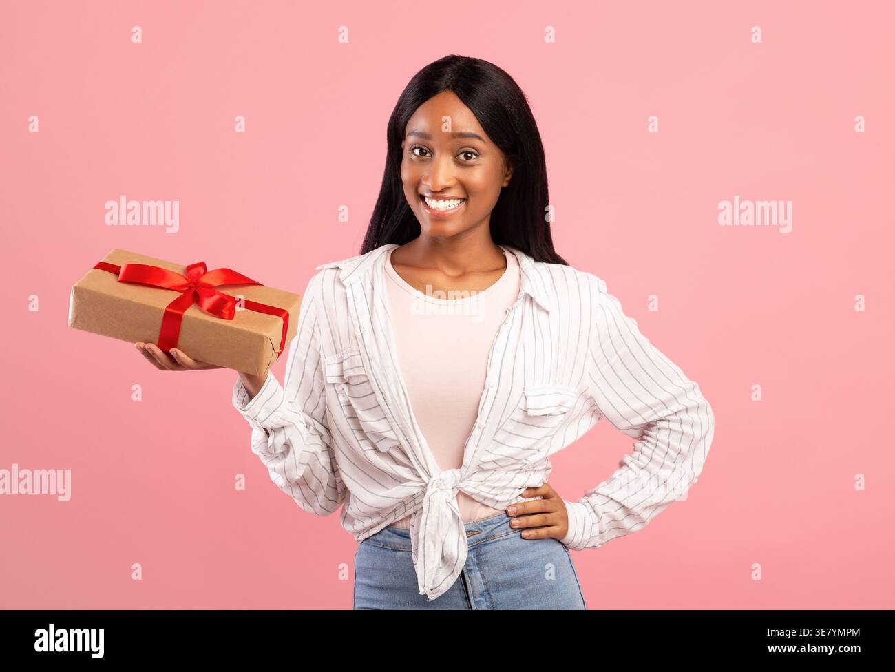 Femme noire souriante avec de longs cheveux droits tenant la boîte-cadeau avec ruban rouge tout en se tenant debout avec la main sur les hanches sur fond rose en studio Banque D'Images