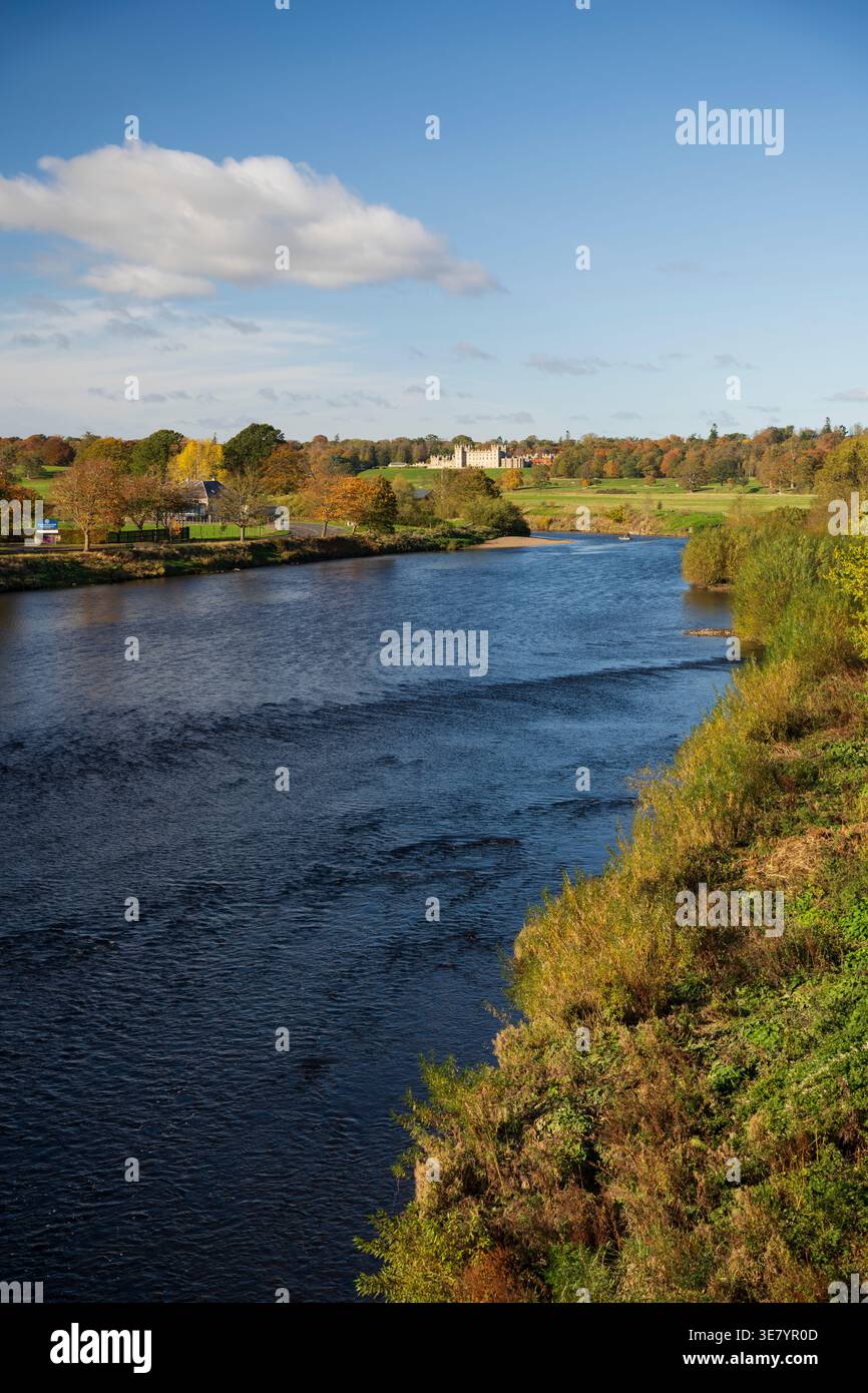 Une large rivière coule à travers un paysage parsemé d'arbres affichant un feuillage d'automne vibrant, menant à un grand domaine sous un ciel bleu avec des nuages Banque D'Images