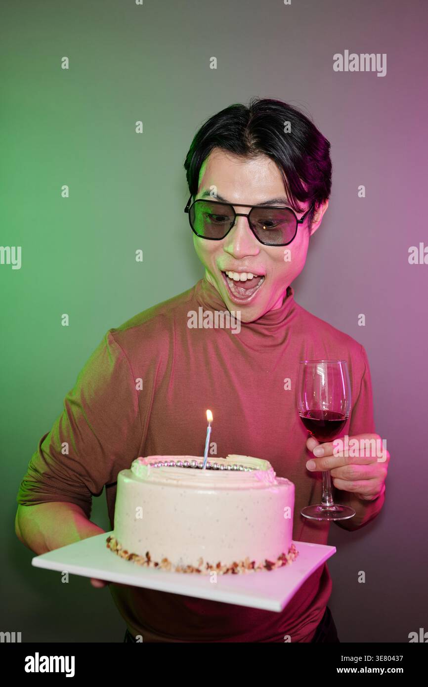 Joyeux homme asiatique portant des lunettes de soleil tenant verre de vin et gâteau avec bougie allumée debout contre le mur avec des néons verts et violets dans St... Banque D'Images