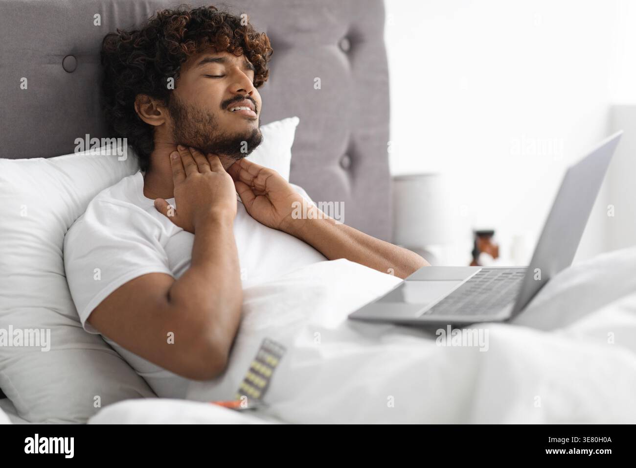 Homme asiatique malade avec les cheveux bouclés touchant le cou couché avec ordinateur portable sur les genoux sur le lit dans la chambre à coucher à la maison Banque D'Images