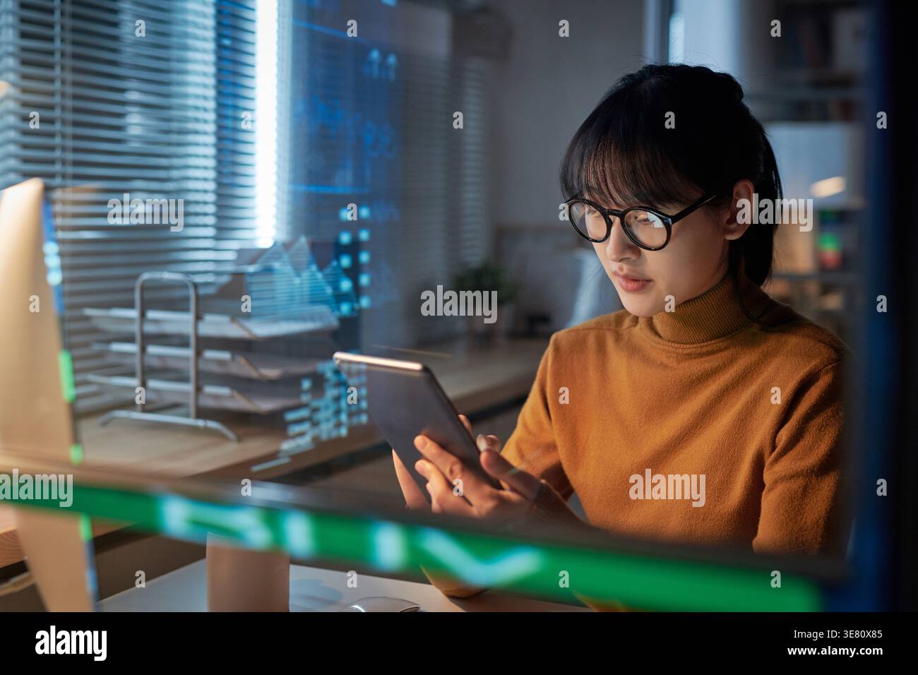 Femme d'affaires asiatique concentrée portant des lunettes en utilisant la tablette assis à table dans le bureau la nuit Banque D'Images