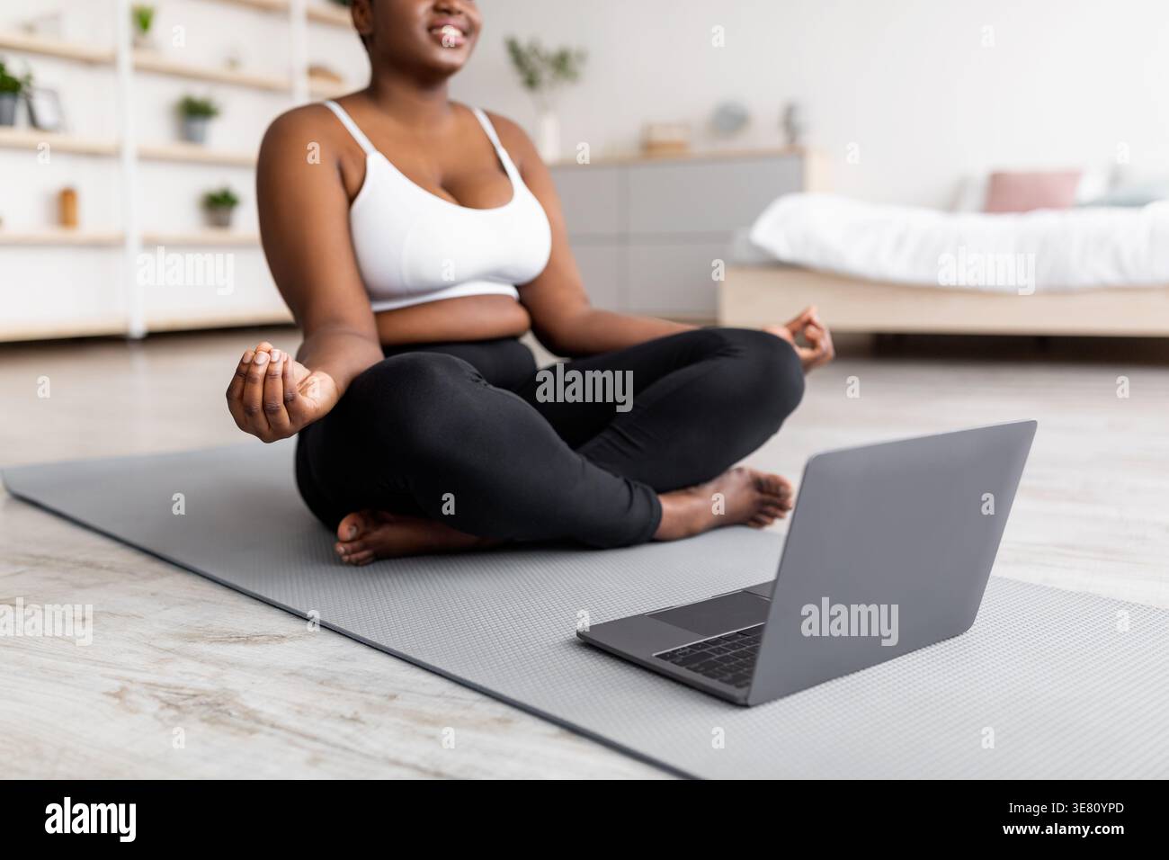 Curvy femme noire assise dans la pose de lotus sur le tapis de yoga près de l'ordinateur portable à la maison Banque D'Images