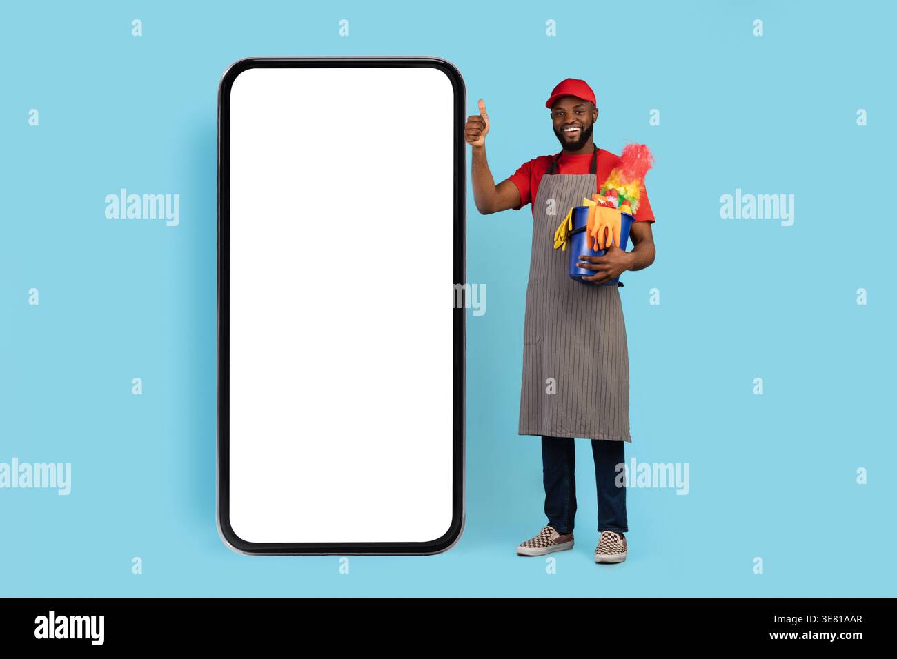 Capuchon de port et tablier de nettoyant noir pour hommes montrant les pouces vers le haut et tenant le panier d'outils de nettoyage debout à côté d'un grand smartphone contre le dos bleu... Banque D'Images