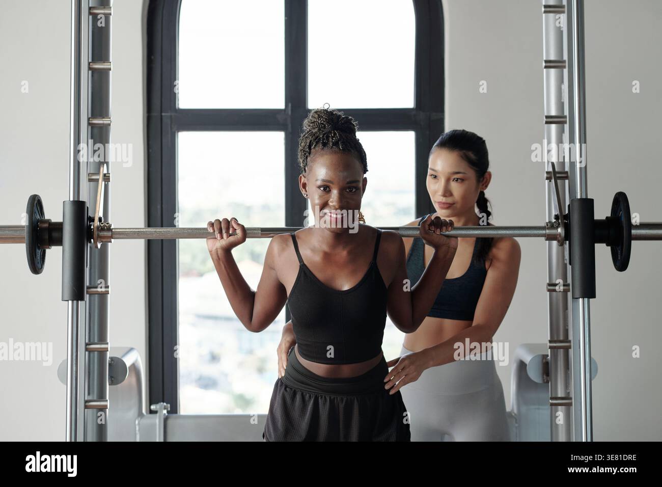 Femme noire se préparant à faire des squats avec barre sur son épaule près de l'entraîneur de fitness féminin asiatique aidant avec sa posture tout en se tenant debout dans le gymnase Banque D'Images