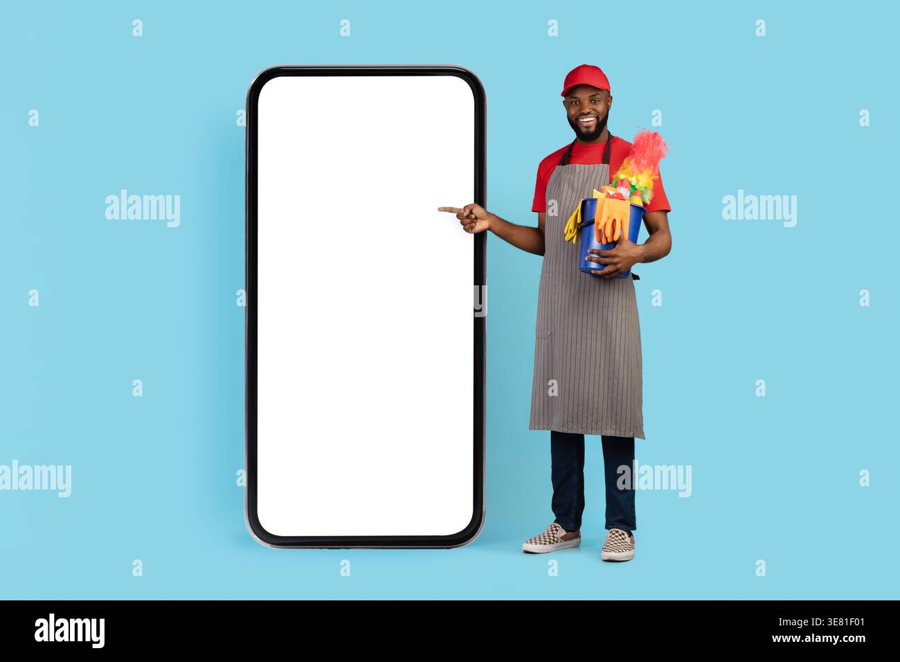 Chapeau de port de nettoyant masculin noir et tablier pointant vers le grand smartphone tenant le panier d'outils de nettoyage debout sur fond bleu dans le studio Banque D'Images