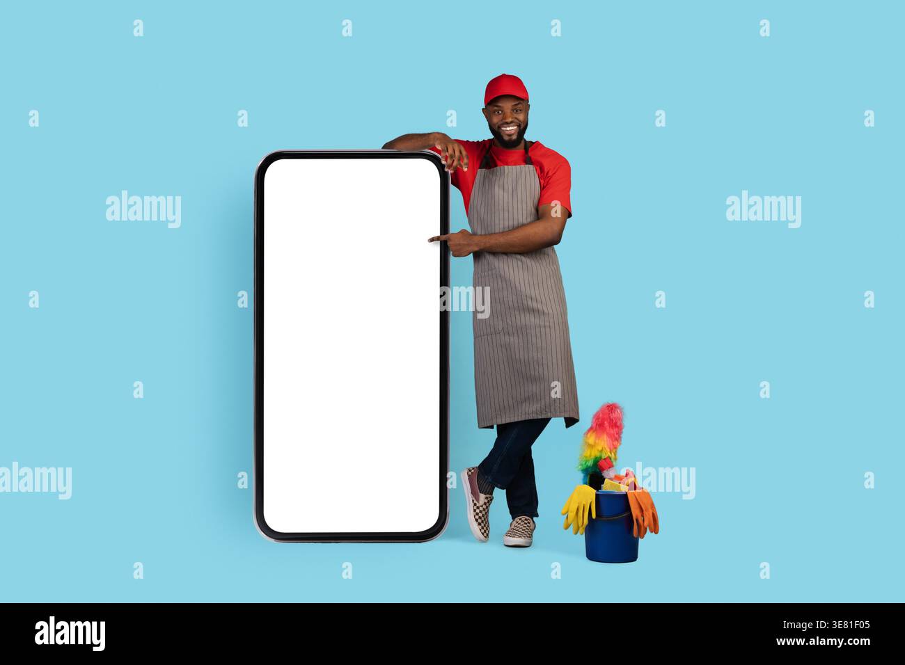 Casquette et tablier noir nettoyant masculin pointant sur le grand smartphone debout avec les jambes croisées sur fond bleu en studio Banque D'Images