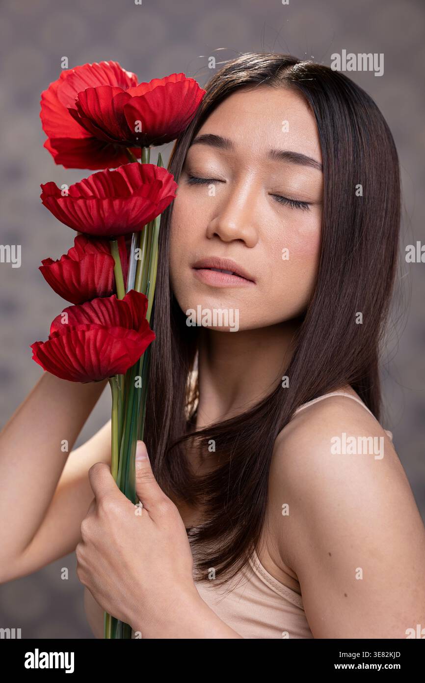 Beau modèle de femme asiatique fermant les yeux à côté du bouquet d'œillets rouges, posant avec une peau lisse brillante pour une publicité de soins de la peau de beauté. Fière posture féminine avec un look naturel. Banque D'Images