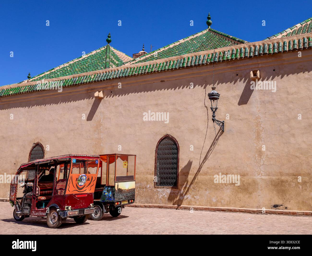 Façade latérale du bâtiment historique de la Madrasa Ben Youssef avec deux tuk-tuks garés à l'extérieur par une journée d'été ensoleillée à Marrakech, Maroc Banque D'Images