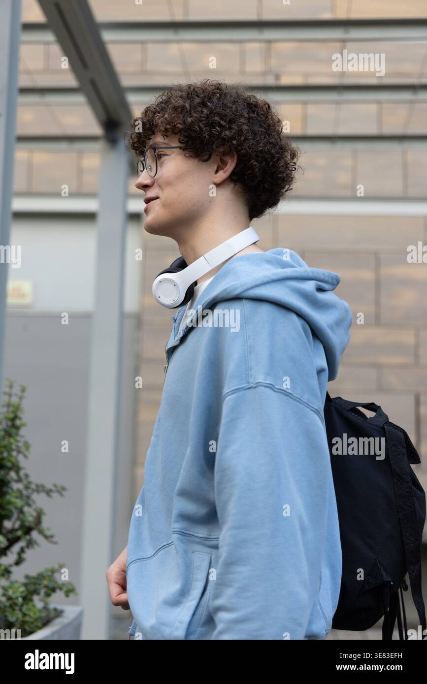 Adolescent se tenant debout regardant à gauche à l'entrée moderne portant un sweat à capuche bleu, des lunettes, des écouteurs Banque D'Images