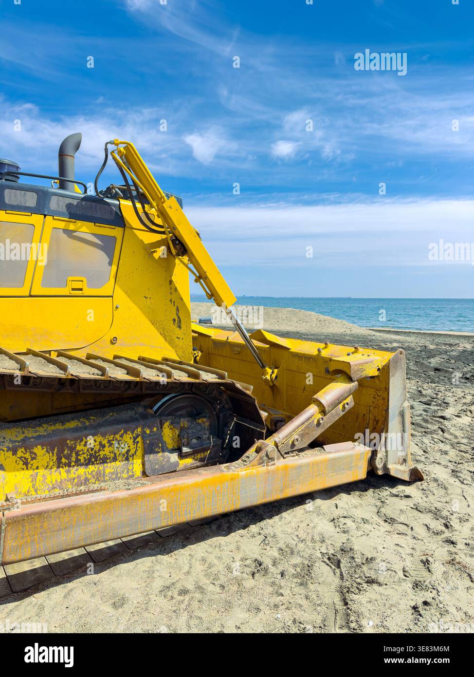 Un bulldozer jaune travaille sur la plage, déplaçant le sable près de l'eau. Le ciel clair et l'océan sont visibles en arrière-plan, montrant l'activité le long des s. Banque D'Images