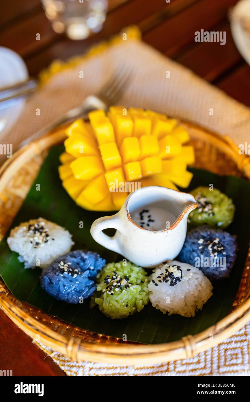 Dessert de riz collant coloré de style thaïlandais avec sauce au lait de mangue et de noix de coco servi sur feuille de banane dans un plateau tissé, gros plan du haut vers le bas sur une table en bois. Banque D'Images