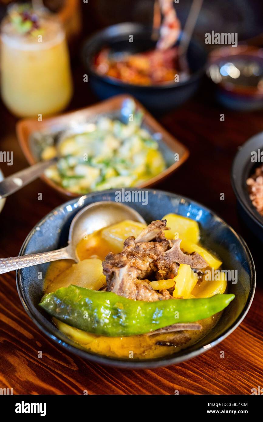 Gros plan d'un ragoût copieux dans un bol bleu avec de la viande, des pommes de terre et un piment vert entier, sur une table en bois avec des plats d'accompagnement flous et boire en b. Banque D'Images