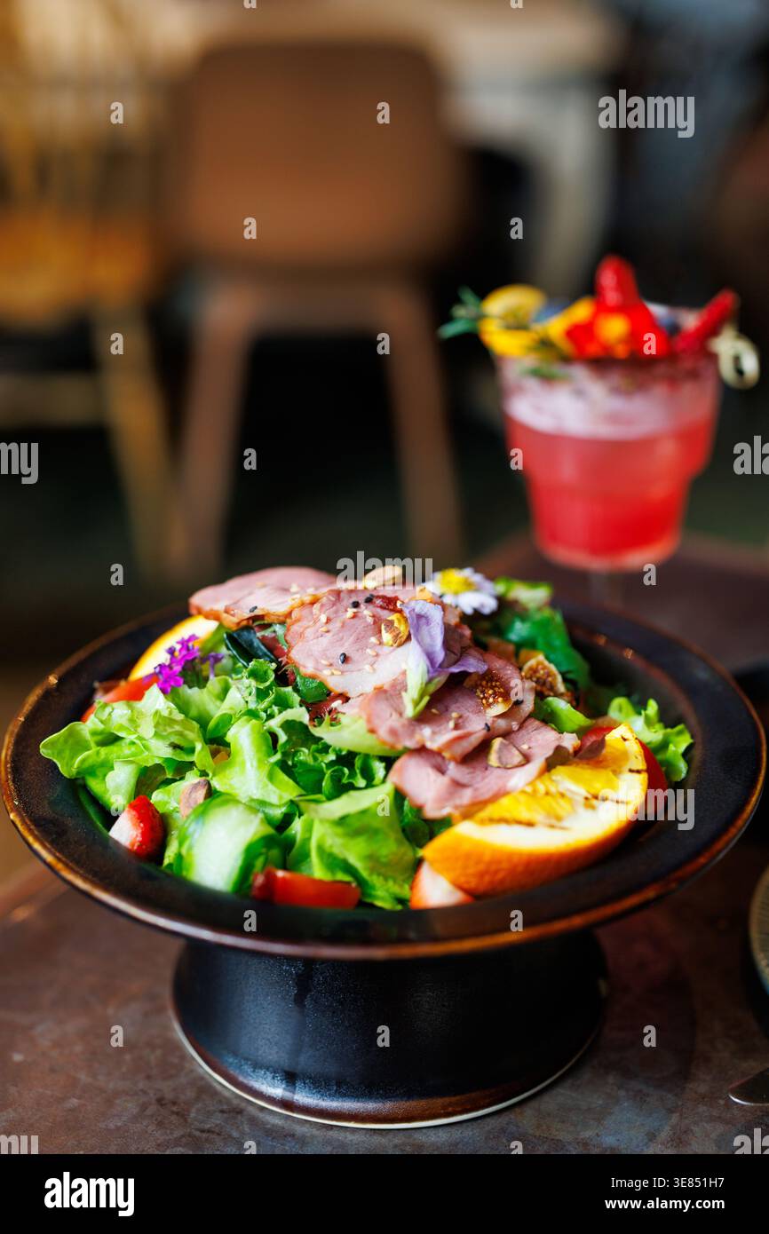 Salade gourmande avec tranches de viande rôtie, légumes verts, fruits et noix dans un bol noir ; fond de café flou avec un cocktail de fruits rouges et paille. Banque D'Images