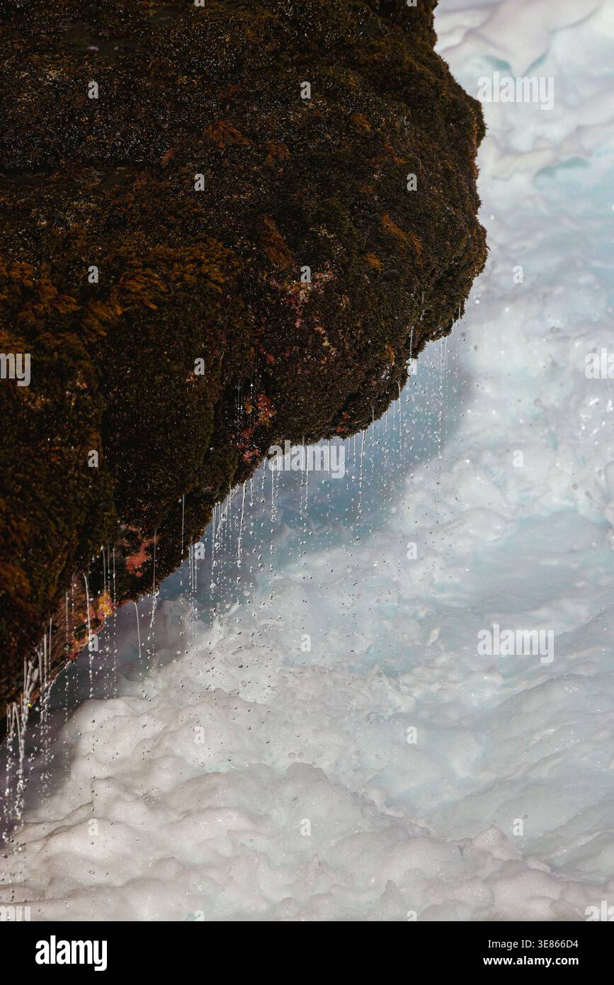 Gros plan d'un surplomb rocheux foncé avec de la mousse et de minces jets d'eau de fonte qui coulent sur une surface glacée bleu pâle et blanc, créant un texte alpin froid Banque D'Images
