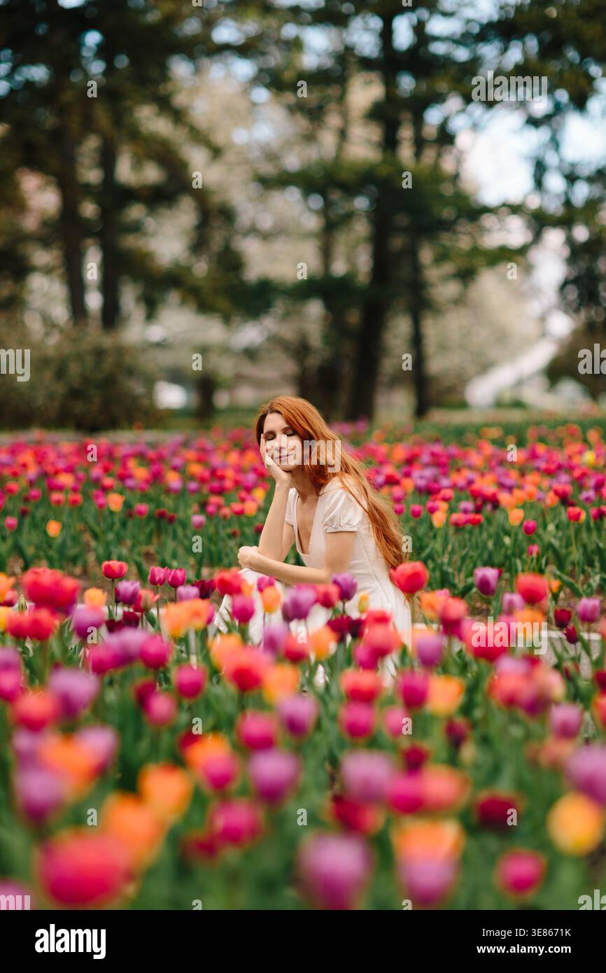 Jeune femme sereine aux cheveux rouges dans une robe blanche reposant parmi les tulipes vibrantes dans un parc de printemps, les arbres doux derrière, l'humeur paisible et la beauté naturelle Banque D'Images