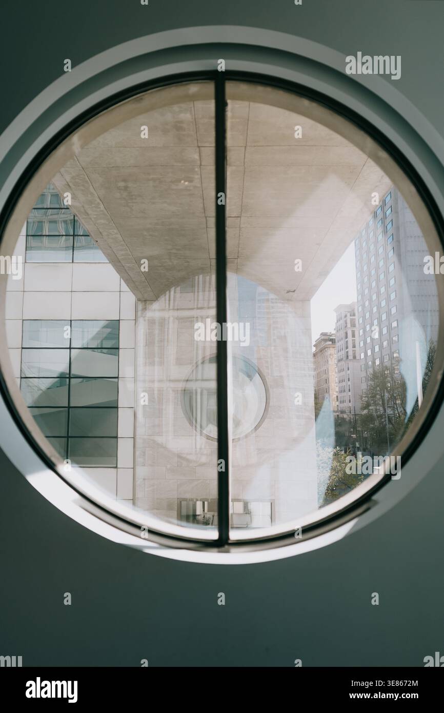 Scène urbaine abstraite vue à travers une fenêtre circulaire, montrant l'architecture moderne en béton et en verre, les lignes géométriques, la lumière du jour douce et le bâtiment de la ville Banque D'Images