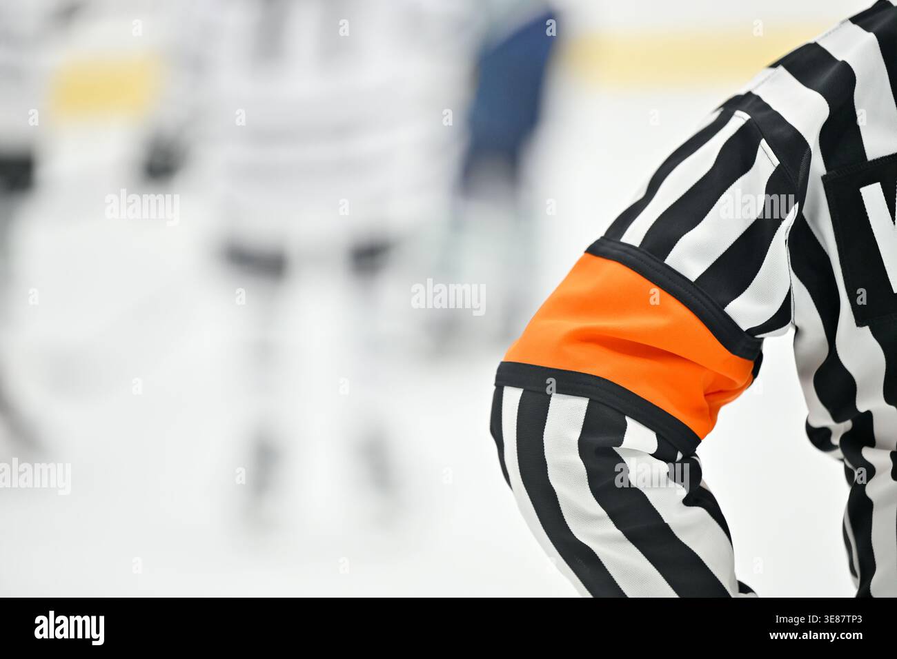 Arbitre de hockey sur glace en uniforme rayé avec brassard orange pendant un match, avec des joueurs flous en arrière-plan sur la patinoire. Banque D'Images