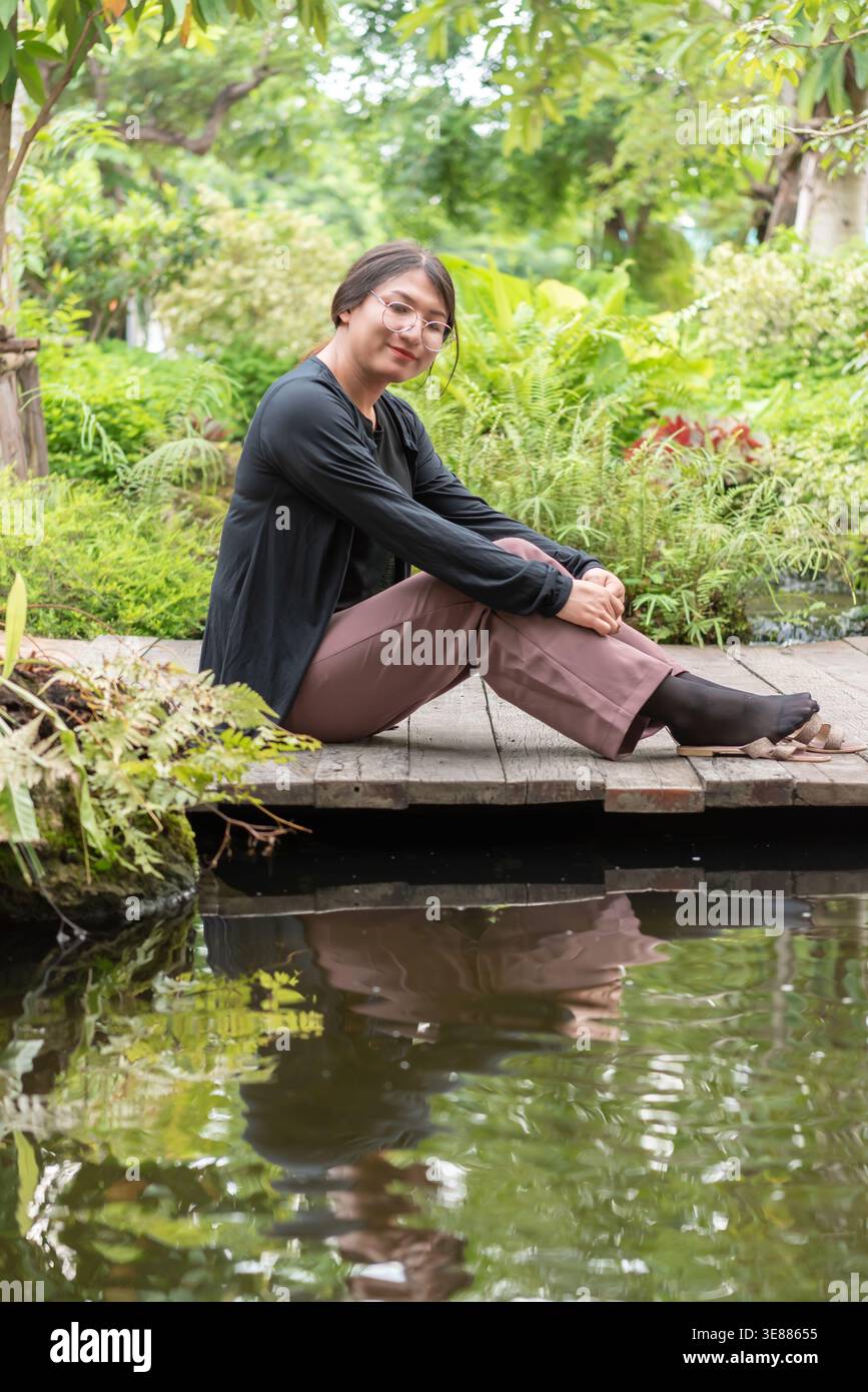 Femme LGBTQ assise près d'un étang dans un jardin luxuriant portant des lunettes détendues pose lumière naturelle cadre extérieur paisible reflétant une identité calme et inclusive Banque D'Images