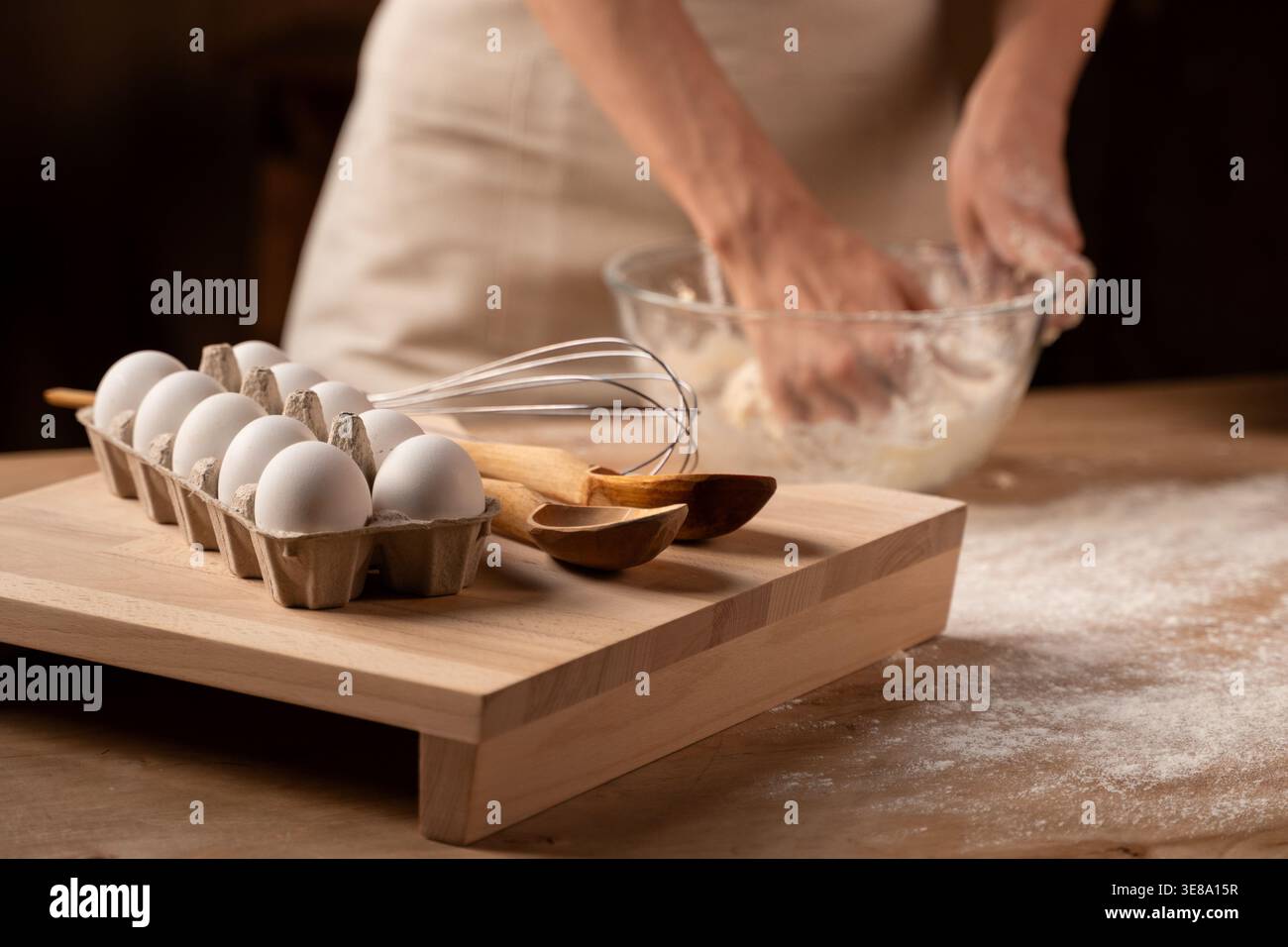 Mains pétrissant la pâte dans un bol en verre, préparant des produits de boulangerie faits maison, des œufs frais et des ustensiles de cuisson sur une planche de bois Banque D'Images