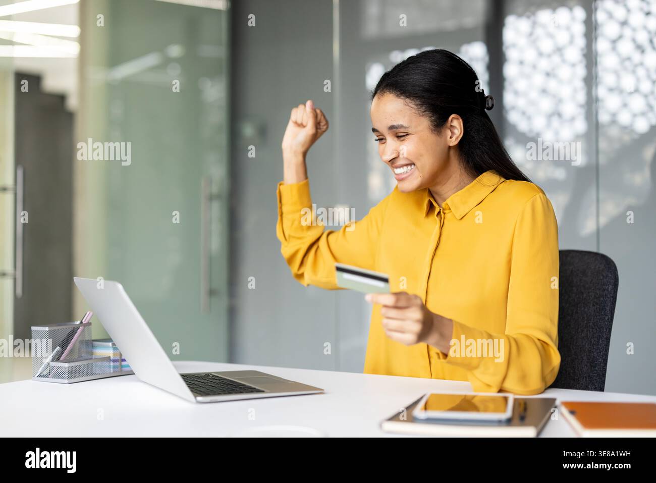 Jeune femme célèbre l'achat en ligne réussi, tenant une carte de crédit et levant le poing tout en travaillant à un ordinateur portable sur un bureau bien rangé, excitée et satisfaite de la transaction Banque D'Images