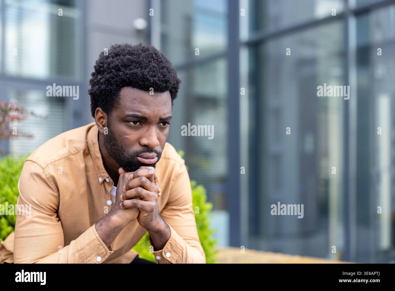 Jeune homme adulte assis à l'extérieur avec les mains serrées, contemplant profondément, exprimant le sérieux et l'inquiétude tout en éprouvant la lutte émotionnelle dans un environnement urbain Banque D'Images
