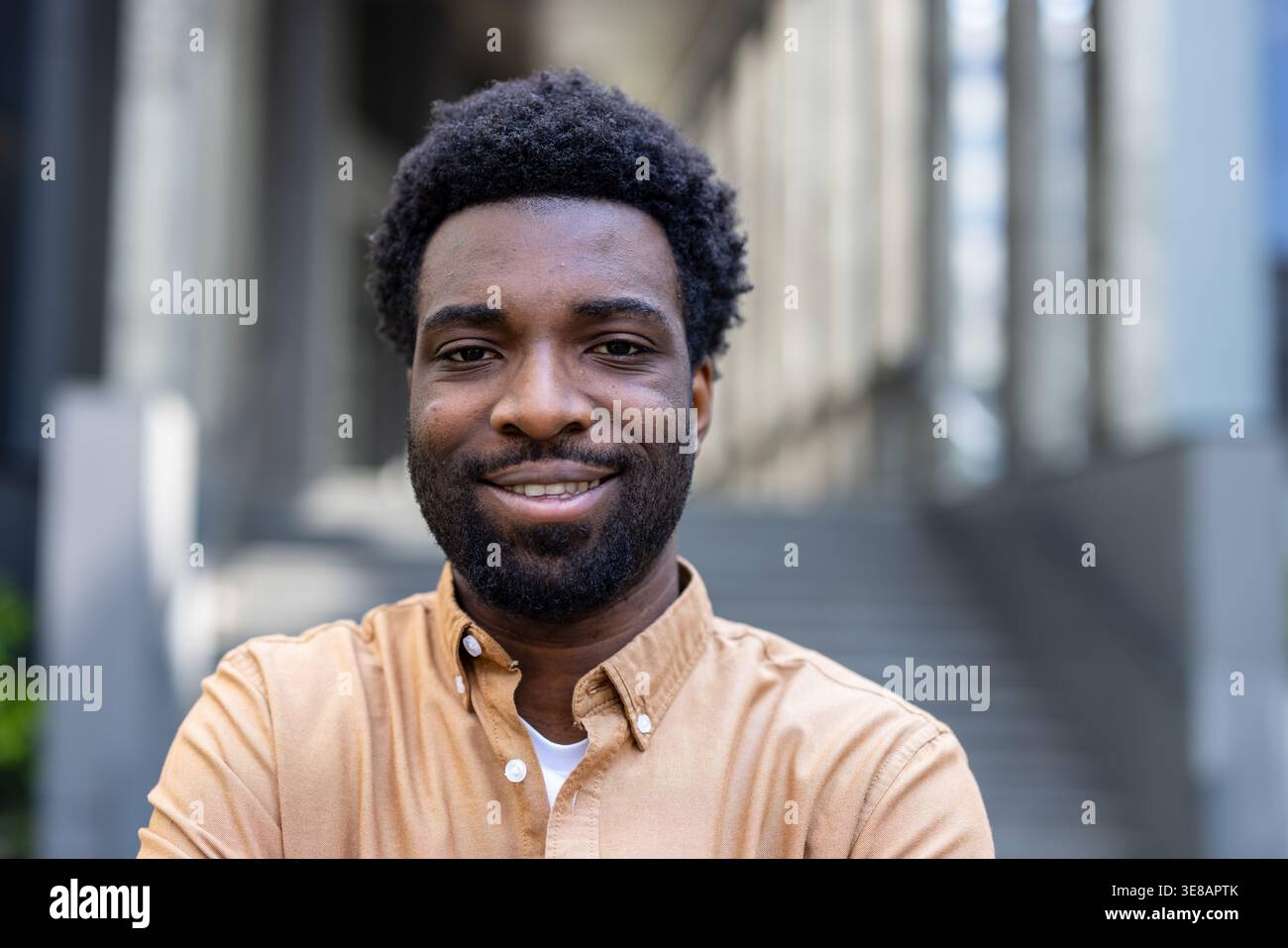 Jeune homme souriant à la caméra avec une expression authentique, véhiculant positivité et succès tout en se tenant dans un environnement urbain contemporain avec des détails architecturaux en arrière-plan Banque D'Images